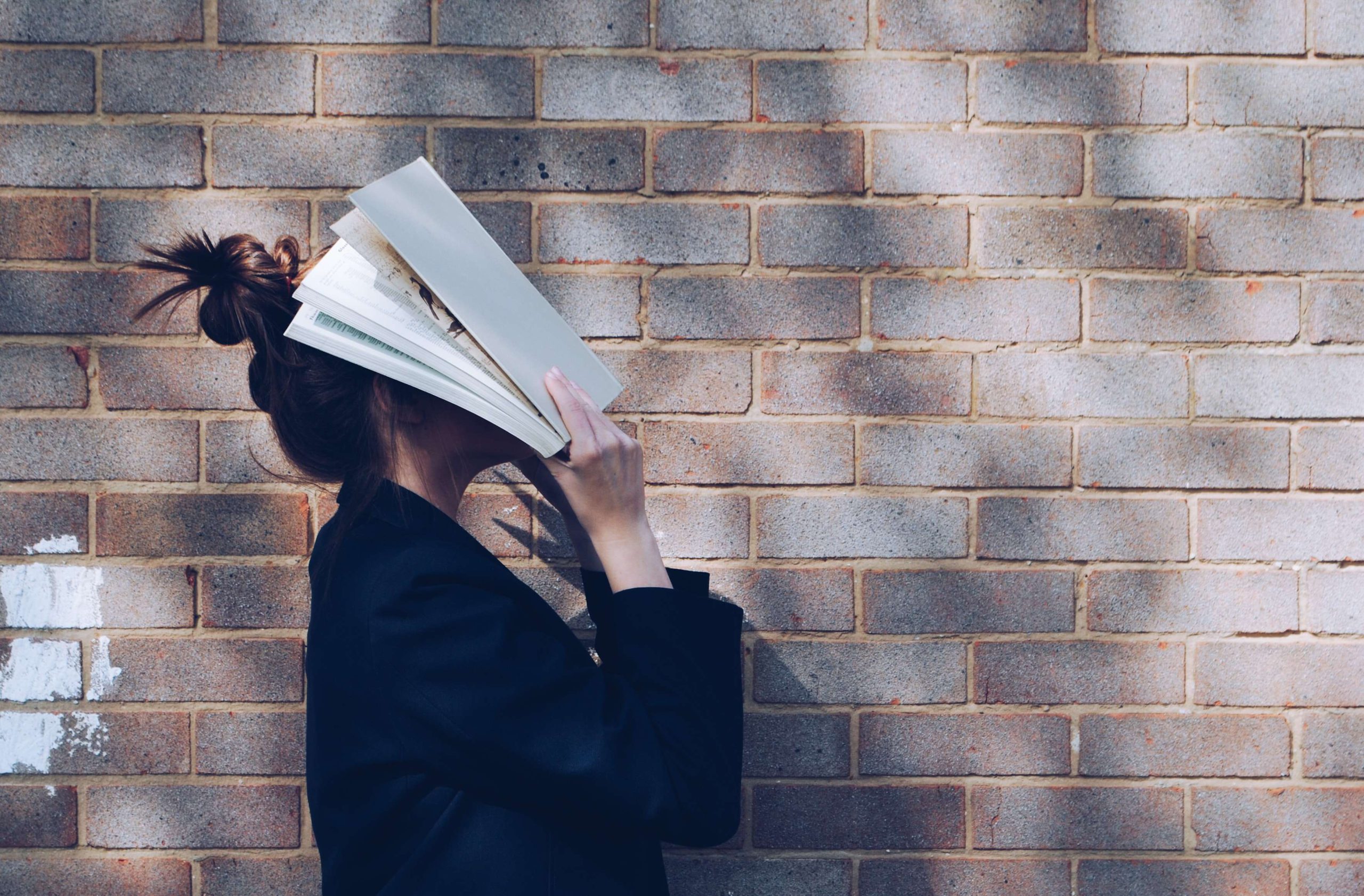 Woman standing next to a brick wall with her head immersed in a book